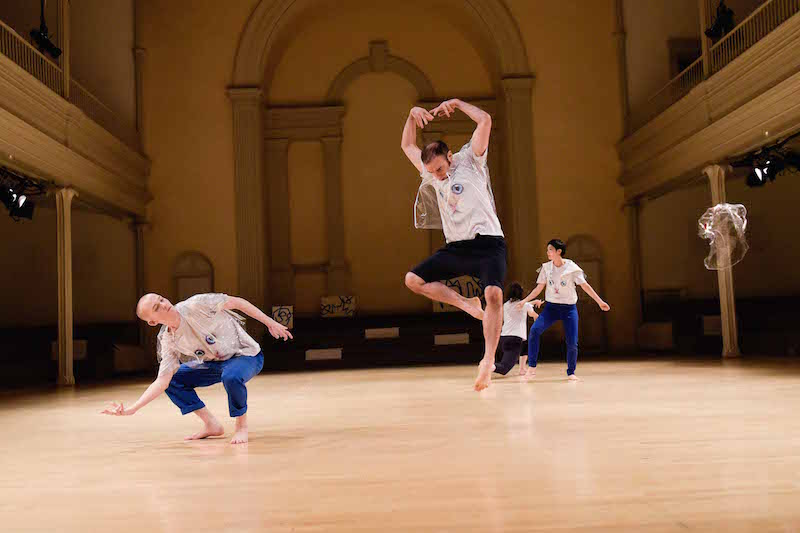 Dancers in cat t-shirts and transparent jackets. A man leaps into the air with feline-like prowess